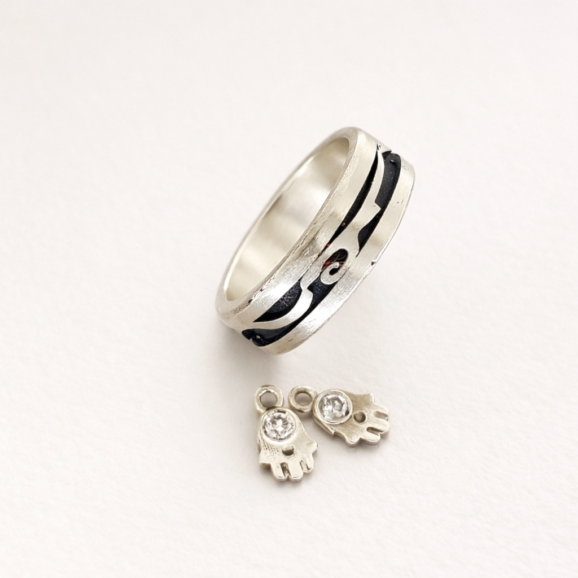 טבעת כסף סטרלינג בעבודת יד עם חריטה כהה ותליוני חמסה על רקע קרם של סטודיו צורפות פלוס