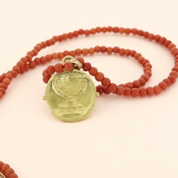 שרשרת אלמוגים אדומים עם תליון זהב בעיצוב מטבע רומי, על רקע שמנת בהיר חם בסגנון צורפות פלוס.